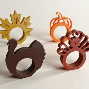 May include: Four wooden napkin rings in fall shapes: a yellow maple leaf, an orange pumpkin, a brown turkey, and a reddish-brown turkey. Each ring has a circular opening for a napkin. Perfect for Thanksgiving or autumn decor.
