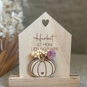Könnte beinhalten: Holzdekoration in Hausform mit Herz-Ausschnitt und Kürbis-Design. Der Text "Herbst ist meine Lieblingsfarbe" ist in das Holz graviert. Der Kürbis hat ein kleines Herzdetail. Trockenblumen sind befestigt.