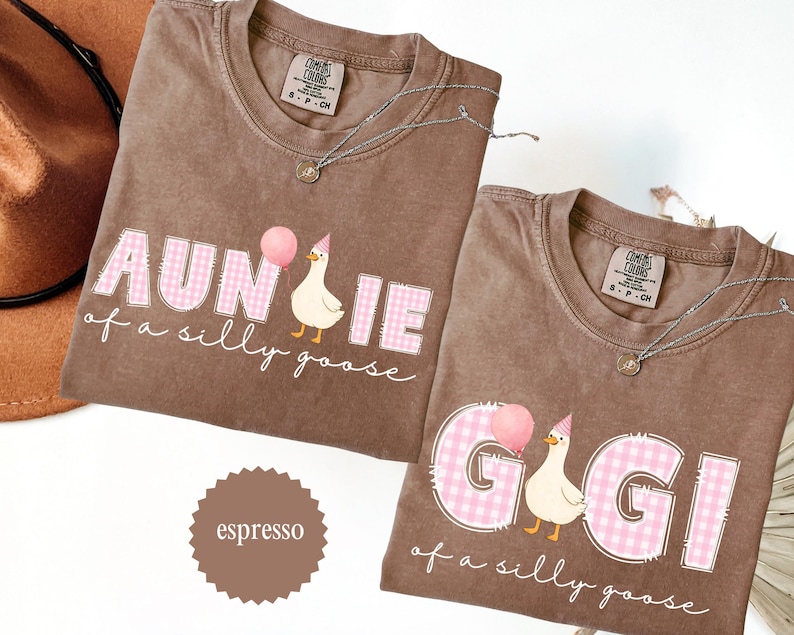 May include: Two espresso-colored t-shirts with the phrases "AUNTIE of a silly goose" and "GIGI of a silly goose" in pink gingham letters. Each shirt features a cartoon goose holding a balloon and wearing a party hat. Silver necklaces are draped on the shirts.