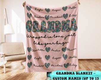 May include: A pink blanket with the word "GRANDMA" in large, floral-patterned letters. The blanket features names and heart shapes. Text reads "Wrapped in love, just like your hugs."