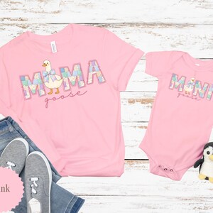 K&ouml;nnte beinhalten: Rosafarbene T-Shirts und ein Baby-Body mit einem "MAMA goose" und "MINI goose" Design. Der Text ist im Patchwork-Stil mit einer Cartoon-Gans-Illustration gehalten. Ein Paar graue Turnschuhe und ein Pinguin-Pl&uuml;schtier sind ebenfalls im Bild.