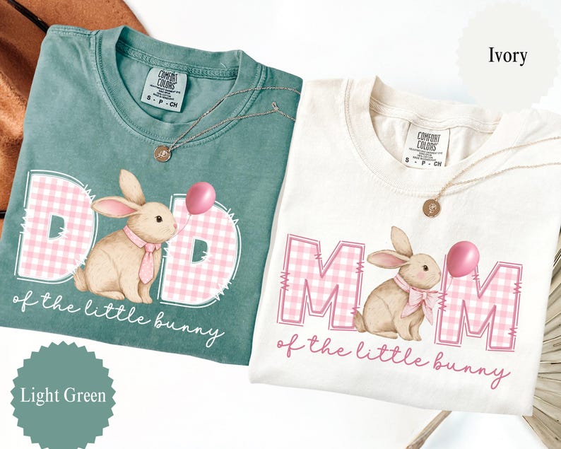 May include: Two t-shirts, one light green and one ivory, with the text "DAD" and "MOM" in pink gingham, and a bunny graphic. The text "of the little bunny" is below the graphics. Each shirt has a Comfort Colors tag.