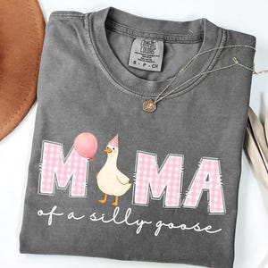 May include: Dark gray Comfort Colors t-shirt with the words "MAMA of a silly goose" in white script. The word "MAMA" is in pink gingham print. A cartoon goose wearing a party hat and holding a pink balloon is featured.