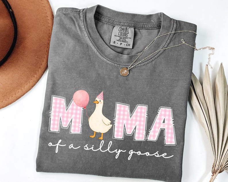 May include: Dark gray Comfort Colors t-shirt with the words "MAMA of a silly goose" in white script. The word "MAMA" is in pink gingham print. A cartoon goose wearing a party hat and holding a pink balloon is featured.