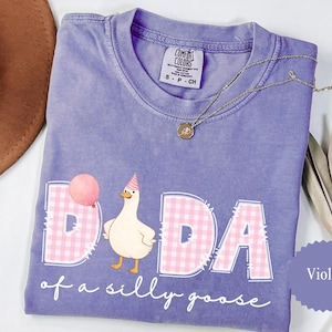 May include: Violet Comfort Colors t-shirt with the phrase "DADA of a silly goose" and a cartoon goose wearing a party hat. The word "DADA" is in pink gingham print. A gold necklace is draped across the shirt.
