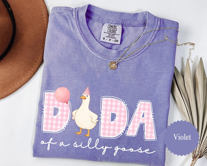 May include: Violet Comfort Colors t-shirt with the phrase "DADA of a silly goose" and a cartoon goose wearing a party hat. The word "DADA" is in pink gingham print. A gold necklace is draped across the shirt.