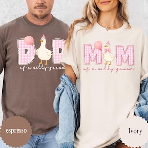 May include: Espresso and ivory-colored t-shirts with matching graphics. The shirts feature the words "DAD" and "MOM" in pink gingham, with a cartoon goose wearing a party hat and holding a balloon. The phrase "of a silly goose" is written below.