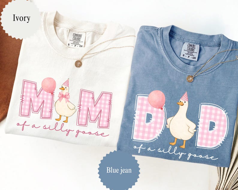 May include: Two t-shirts, one ivory and one blue jean, with the words "MOM" and "DAD" in pink gingham. Each shirt features a cartoon goose holding a balloon and wearing a party hat, with the phrase "of a silly goose". Gold necklaces are draped on the shirts.