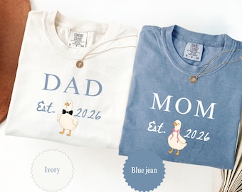 May include: Two Comfort Colors t-shirts, one ivory and one blue jean, with gold necklaces. The ivory shirt says "DAD Est. 2026" with a duck graphic. The blue jean shirt says "MOM Est. 2026" with a duck graphic.