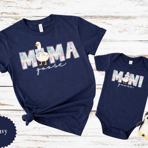 K&ouml;nnte beinhalten: Marineblaues Set aus passenden T-Shirts und Bodys. Das Erwachsenen-Shirt tr&auml;gt die Aufschrift "MAMA goose" mit einer G&auml;nsegrafik. Der Baby-Body tr&auml;gt die Aufschrift "MINI goose" mit einer passenden G&auml;nsegrafik. Ein kleines Pinguin-Spielzeug liegt neben dem Body.