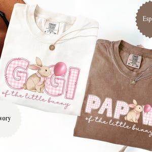 May include: Two Comfort Colors t-shirts, one ivory and one espresso, featuring the text "Gigi" and "Papaw" with a bunny graphic and the phrase "of the little bunny". Each shirt has a round gold pendant necklace.