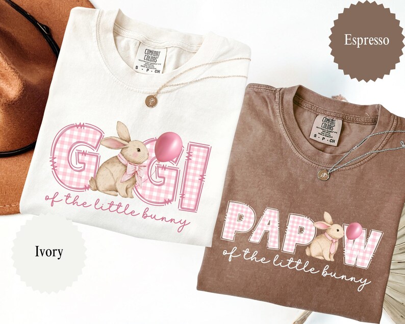 May include: Two Comfort Colors t-shirts, one ivory and one espresso, featuring the text "Gigi" and "Papaw" with a bunny graphic and the phrase "of the little bunny". Each shirt has a round gold pendant necklace.