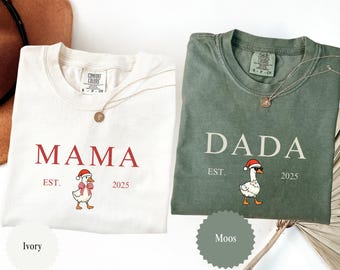 May include: Two Comfort Colors t-shirts, ivory and moss green, with the words "MAMA" and "DADA" printed in red and white, respectively. Each shirt features a cartoon goose wearing a Santa hat and the year "2025". A gold necklace is draped over each shirt.