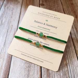 Pode incluir: Duas pulseiras de macramé verde com contas de aventurina verde e detalhes dourados são exibidas em um cartão. O cartão diz "Balance & Harmony" e "Green Aventurine". As pulseiras são feitas à mão.