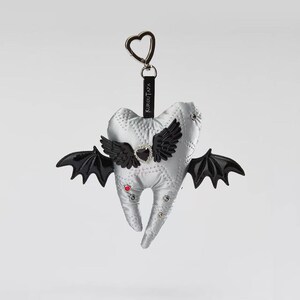 Puede incluir: Un llavero de peluche con forma de diente plateado, alas de murci&eacute;lago negras, un coraz&oacute;n central negro con borde de pedrer&iacute;a y peque&ntilde;os remaches rojos y transparentes. Una etiqueta negra con el texto KUROUTAYA cuelga de un cierre met&aacute;lico.
