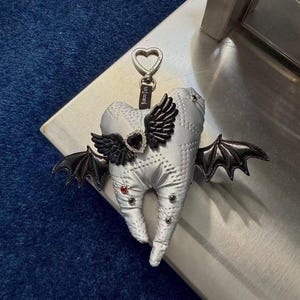 Puede incluir: Un llavero de peluche plateado con forma de diente que presenta alas de murci&eacute;lago negras, un dije central en forma de coraz&oacute;n negro con borde de pedrer&iacute;a y peque&ntilde;os remaches rojos y negros. Un cierre plateado en forma de coraz&oacute;n lleva una etiqueta negra KIROUTAYA.