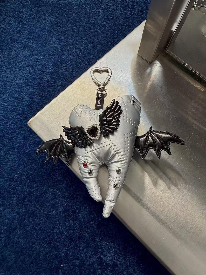 Puede incluir: Un llavero de peluche plateado con forma de diente que presenta alas de murci&eacute;lago negras, un dije central en forma de coraz&oacute;n negro con borde de pedrer&iacute;a y peque&ntilde;os remaches rojos y negros. Un cierre plateado en forma de coraz&oacute;n lleva una etiqueta negra KIROUTAYA.