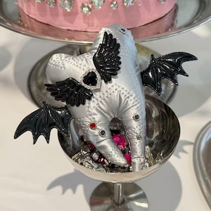 Puede incluir: Un adorno de peluche plateado con forma de diente que presenta alas de murci&eacute;lago negras, un detalle de piedra preciosa negra en forma de coraz&oacute;n y peque&ntilde;os tachones de colores. El peluche descansa en una copa de metal plateado con gemas.