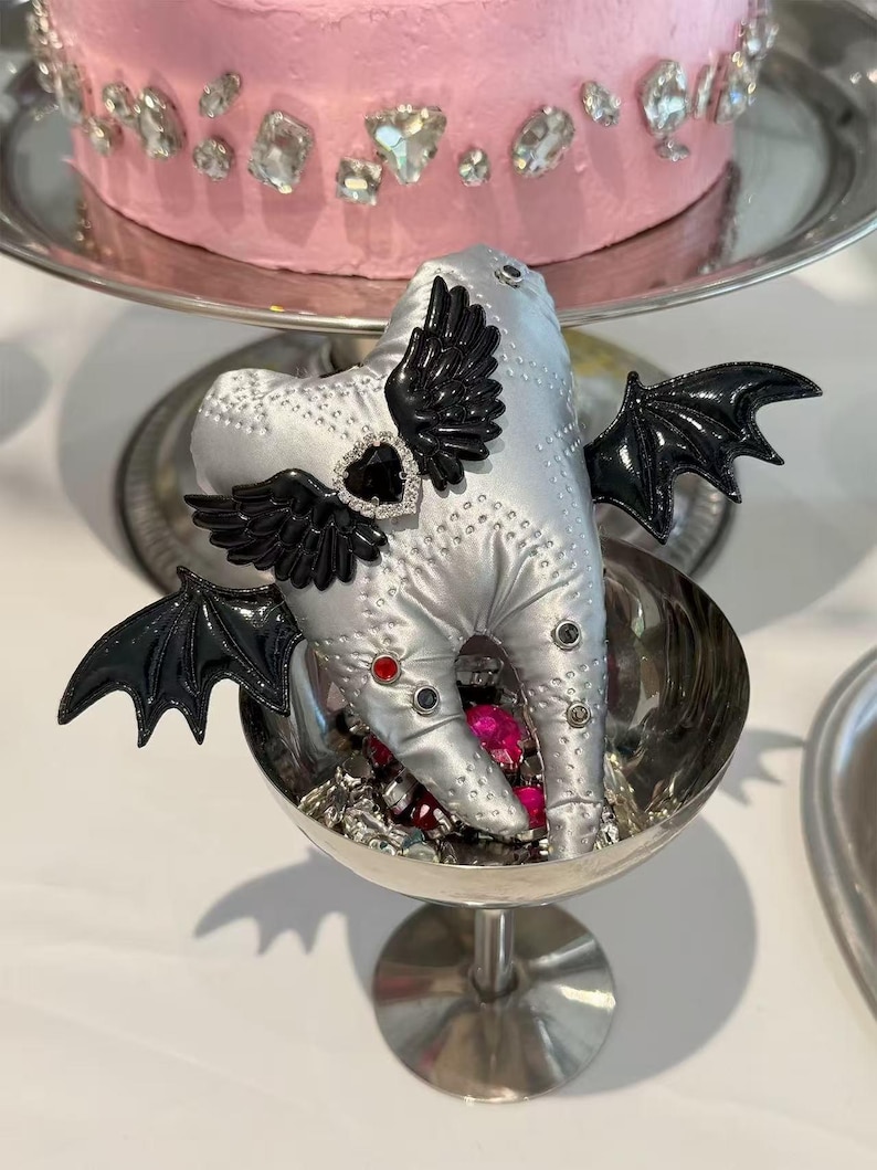 Puede incluir: Un adorno de peluche plateado con forma de diente que presenta alas de murci&eacute;lago negras, un detalle de piedra preciosa negra en forma de coraz&oacute;n y peque&ntilde;os tachones de colores. El peluche descansa en una copa de metal plateado con gemas.
