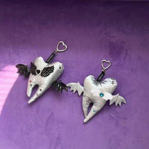 Puede incluir: Dos llaveros de peluche plateados con forma de diente, acolchados y con anillas en forma de coraz&oacute;n. Uno tiene alas de murci&eacute;lago negras y un coraz&oacute;n negro, el otro alas blancas y un coraz&oacute;n azul. Ambos tienen peque&ntilde;os brillantes de colores.