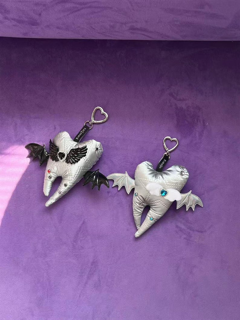 Puede incluir: Dos llaveros de peluche plateados con forma de diente, acolchados y con anillas en forma de coraz&oacute;n. Uno tiene alas de murci&eacute;lago negras y un coraz&oacute;n negro, el otro alas blancas y un coraz&oacute;n azul. Ambos tienen peque&ntilde;os brillantes de colores.