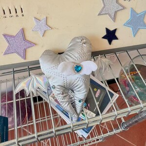 Puede incluir: Un peluche plateado acolchado con forma de diente que presenta alas de murci&eacute;lago iridiscentes, peque&ntilde;os diamantes de imitaci&oacute;n circulares y un detalle central de fieltro blanco con una gema azul en forma de coraz&oacute;n sobre una rejilla met&aacute;lica.
