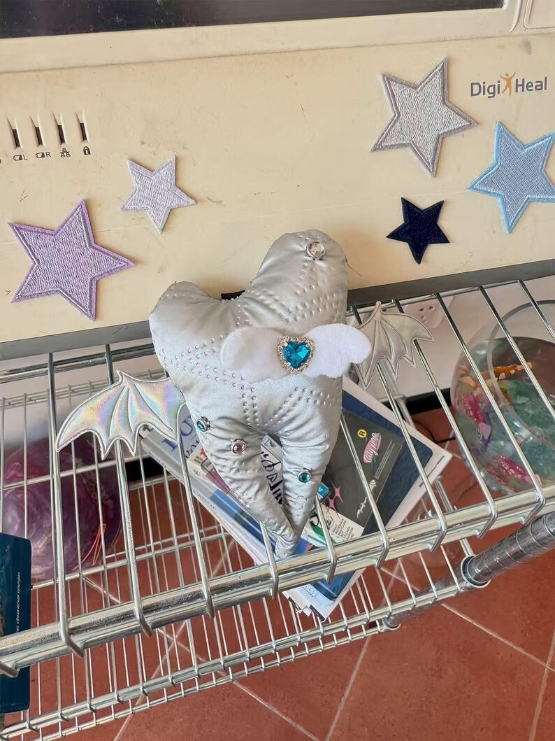 Puede incluir: Un peluche plateado acolchado con forma de diente que presenta alas de murci&eacute;lago iridiscentes, peque&ntilde;os diamantes de imitaci&oacute;n circulares y un detalle central de fieltro blanco con una gema azul en forma de coraz&oacute;n sobre una rejilla met&aacute;lica.