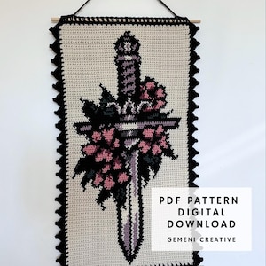 May include: Crocheted wall hanging featuring a sword design with pink flowers and black leaves. The artwork is bordered with a black scalloped edge and includes the text "PDF PATTERN DIGITAL DOWNLOAD" and "GEMENI CREATIVE".