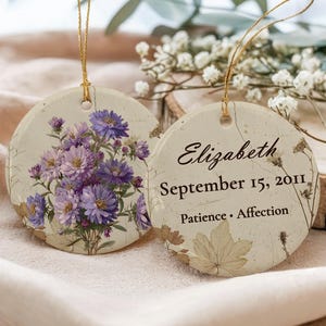 May include: Two round ceramic ornaments with gold hanging strings. One features a cluster of purple aster flowers on a cream background. The other displays the text Elizabeth, September 15, 2011, and Patience • Affection, accented with botanical illustrations.