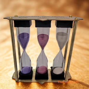 以下が含まれることがあります： 真鍮色のティータイマー。3つの砂時計があり、それぞれ紫、ピンク、白の砂が入っています。「PERFECT TEA TIMER」の文字と、3分、4分、5分の時間が表示されています。
