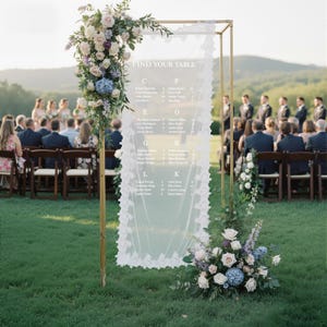 May include: A gold metal frame holds a sheer white fabric with a lace trim and the words "FIND YOUR TABLE". The fabric displays guest names. Floral arrangements of white, blue, and purple flowers adorn the frame and base, set on a green lawn.