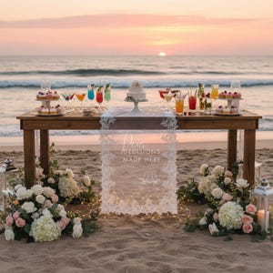 May include: A wooden table on a beach featuring a white lace runner with the text "Pour DECISIONS MADE HERE" and a line drawing of two champagne flutes. The table holds a small white cake, colourful cocktails, and cupcakes at sunset.