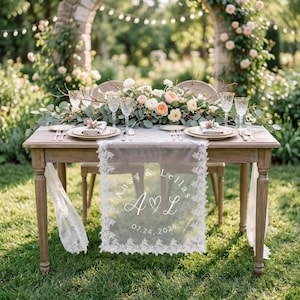 May include: A wooden table set for two with a white lace table runner featuring the printed text Anas & Lellas, the initials A and L with a heart, and the date 07.24.2026. The table includes plates, glassware, and a floral centerpiece.