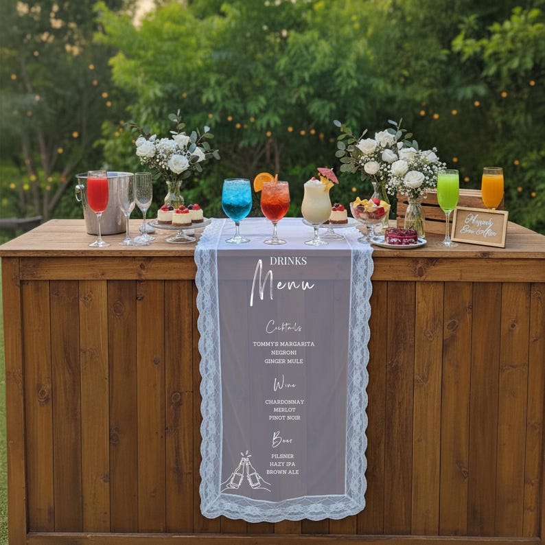 May include: A wooden bar table draped with a white lace-trimmed runner featuring the text Drinks Menu, Cocktails, Wine, and Beer. The table holds various colourful cocktails in glassware, small desserts, and two vases of white flowers.
