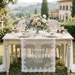 May include: A white lace table runner featuring floral embroidery along the edges and the printed text MR.&MRS. BENNETT est. 2026. The runner is draped over a wooden table set for two with floral china, crystal glassware, and a centerpiece of roses.