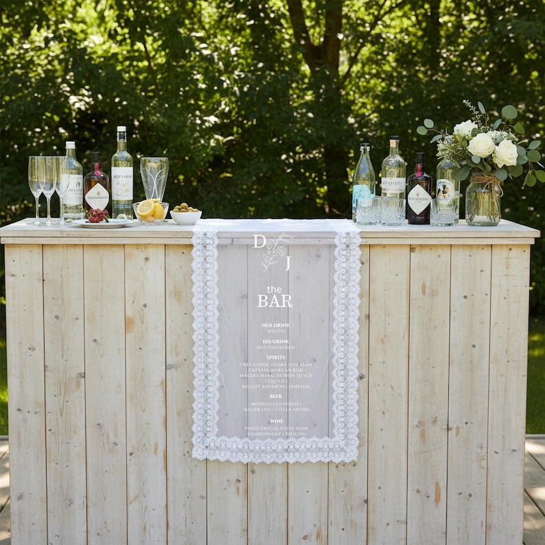 May include: A light-coloured wooden bar featuring a central vertical sign with white lace trim. The sign displays the text D J, the bar, and a drink menu. Several glass bottles, clear drinking glasses, and a small bowl of lemons are on the bar top.