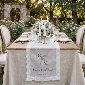 May include: A rectangular table runner made of sheer white fabric with intricate lace trim edges. The center features a black printed design with the initials C and M, the names Casey & Mitchell, and the text est.2026, centered on a wooden table.