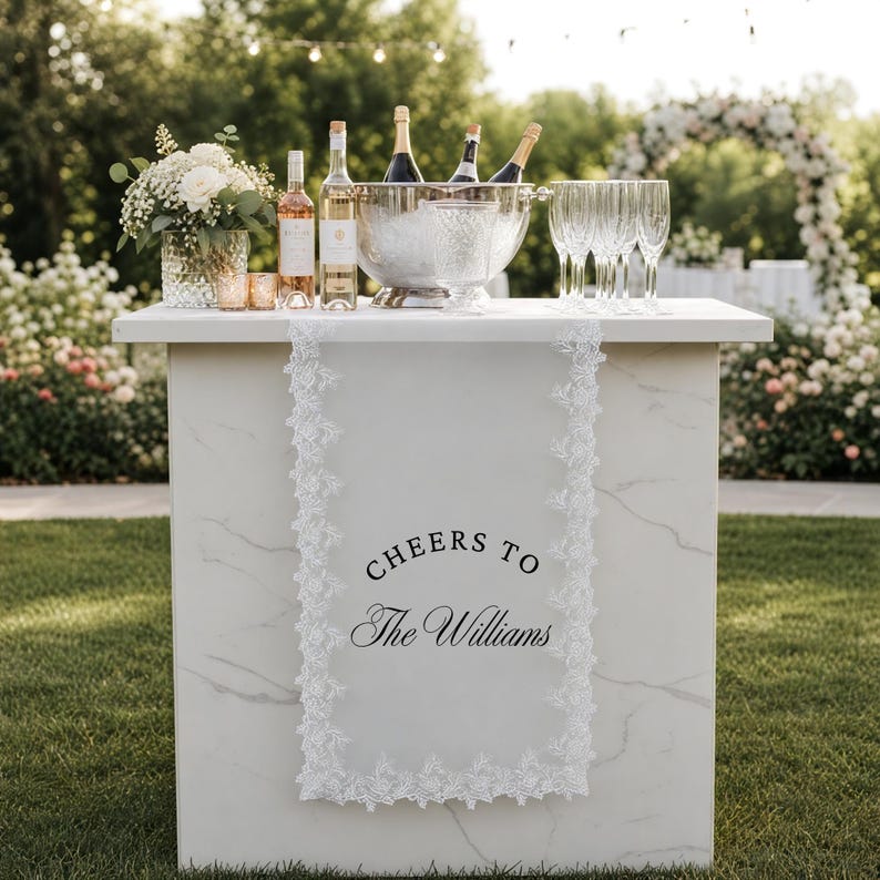 May include: A white table runner with intricate lace edges draped over a marble-patterned bar. The runner features the black printed text "CHEERS TO The Williams" centred on the fabric. The bar is topped with wine bottles, glasses, and flowers.