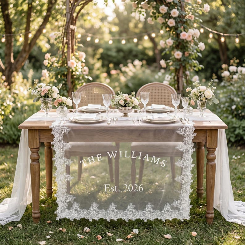 May include: A wooden table set for two with a white lace table runner featuring the text THE WILLIAMS Est. 2026. The table includes white plates, clear glassware, and floral centerpieces, positioned outdoors with two chairs and a floral arch.