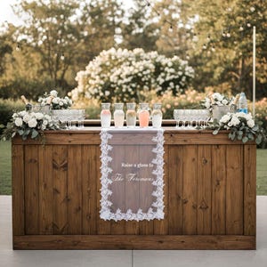 May include: A rectangular wooden bar with vertical planks, topped with five glass carafes containing various coloured liquids, rows of clear glasses, and white floral arrangements. A white lace runner hangs down the front with the text Raise a glass to The Foremans.