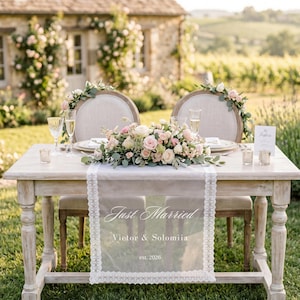 May include: A wooden sweetheart table with a white lace runner featuring the text Just Married, Victor & Solomiia, est. 2026. The table holds a floral centerpiece of pink and white roses, two champagne flutes, and two small glass votive holders.