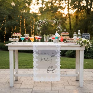 May include: A white wooden table featuring a lace-trimmed runner with the text 'CHEERS TO The Martinez' and a graphic of two clinking champagne flutes. The table is topped with various colourful cocktails, tiered dessert stands, and bottles.