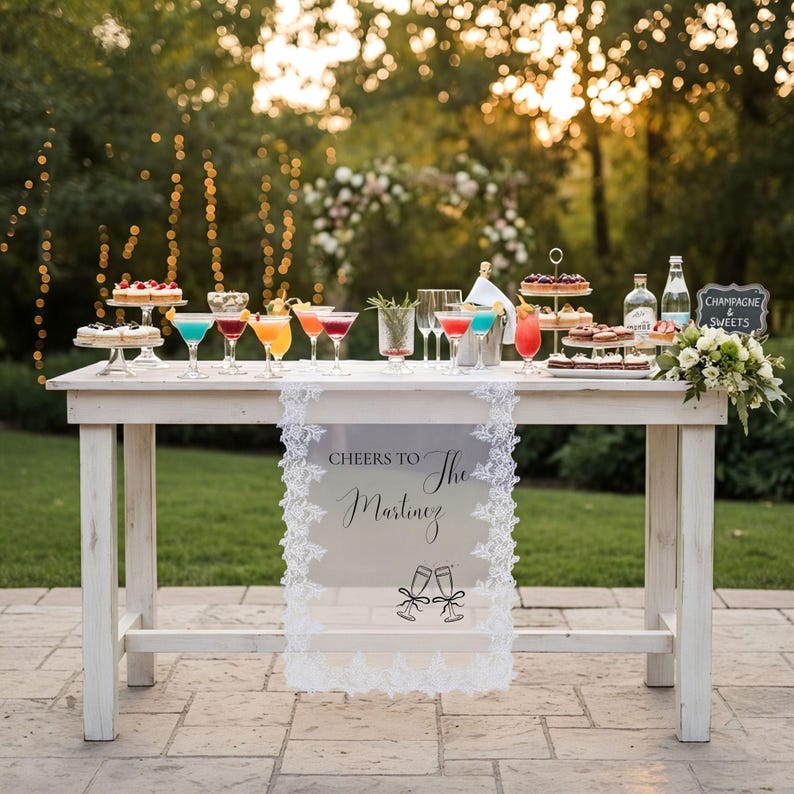 May include: A white wooden table featuring a lace-trimmed runner with the text 'CHEERS TO The Martinez' and a graphic of two clinking champagne flutes. The table is topped with various colourful cocktails, tiered dessert stands, and bottles.