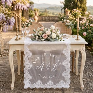 May include: A white lace table runner featuring a scalloped edge and a central printed design with the names Anas & Lellas, the initials A and L separated by a heart, and the date 07.24.2026, draped over a cream-colored wooden table.