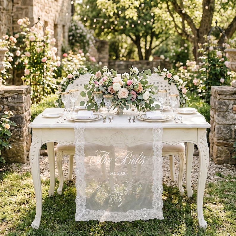 May include: A white wooden table with curved legs features a lace runner printed with the text The Belts est. 2026 and a floral centerpiece of pink and white roses. Two white chairs with cane backs are positioned at the table with place settings.
