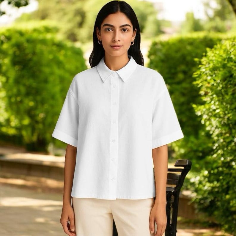 May include: A white short-sleeved shirt with a collar and buttons. The shirt is worn with beige trousers. The background shows green foliage and a park bench.