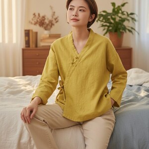 May include: Mustard yellow wrap top with long sleeves and a V-neck. The top is paired with tan pants and white sneakers. The outfit is casual and comfortable, suitable for everyday wear. The top features a tie closure on the side.