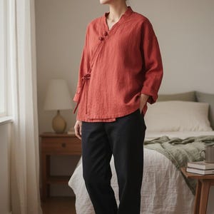 May include: A woman wearing a rust-colored wrap top with three-quarter sleeves and black pants. The top has a V-neck and decorative buttons. The pants are straight-legged. She is wearing white sneakers.