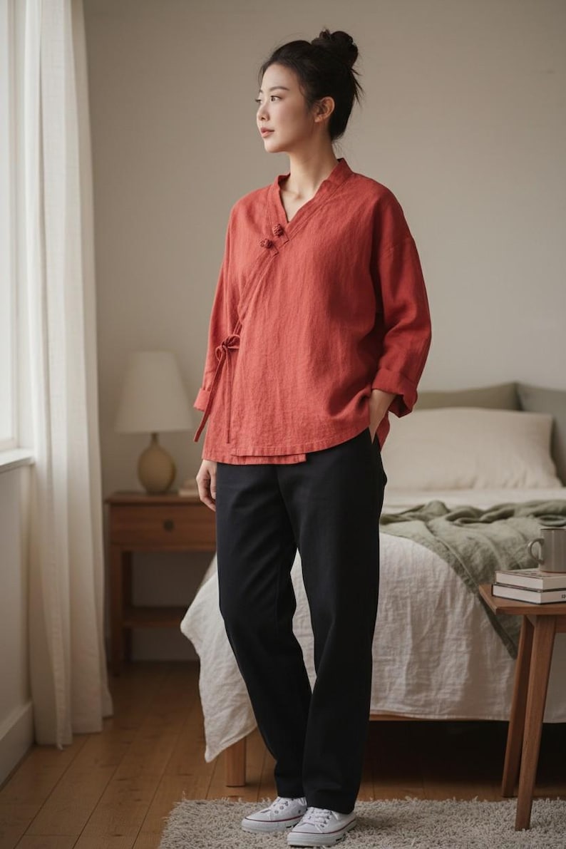 May include: A woman wearing a rust-colored wrap top with three-quarter sleeves and black pants. The top has a V-neck and decorative buttons. The pants are straight-legged. She is wearing white sneakers.