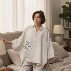 May include: A light-colored linen pajama set. The top is a loose-fitting, button-down shirt with a floral embroidery design in shades of blue and white. The pants are a matching, relaxed fit. The set is displayed in a bedroom setting.